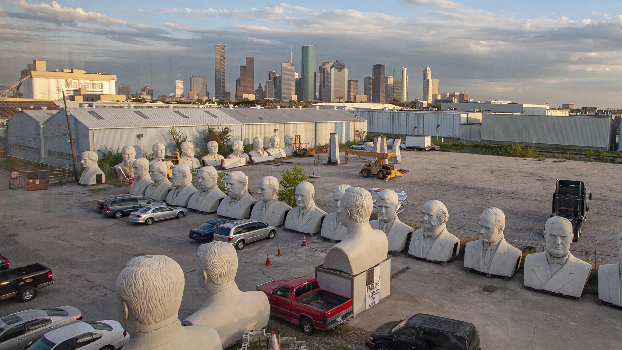 The Houston skyline at dusk behind a lot of monumental sculpted presidential heads in rows